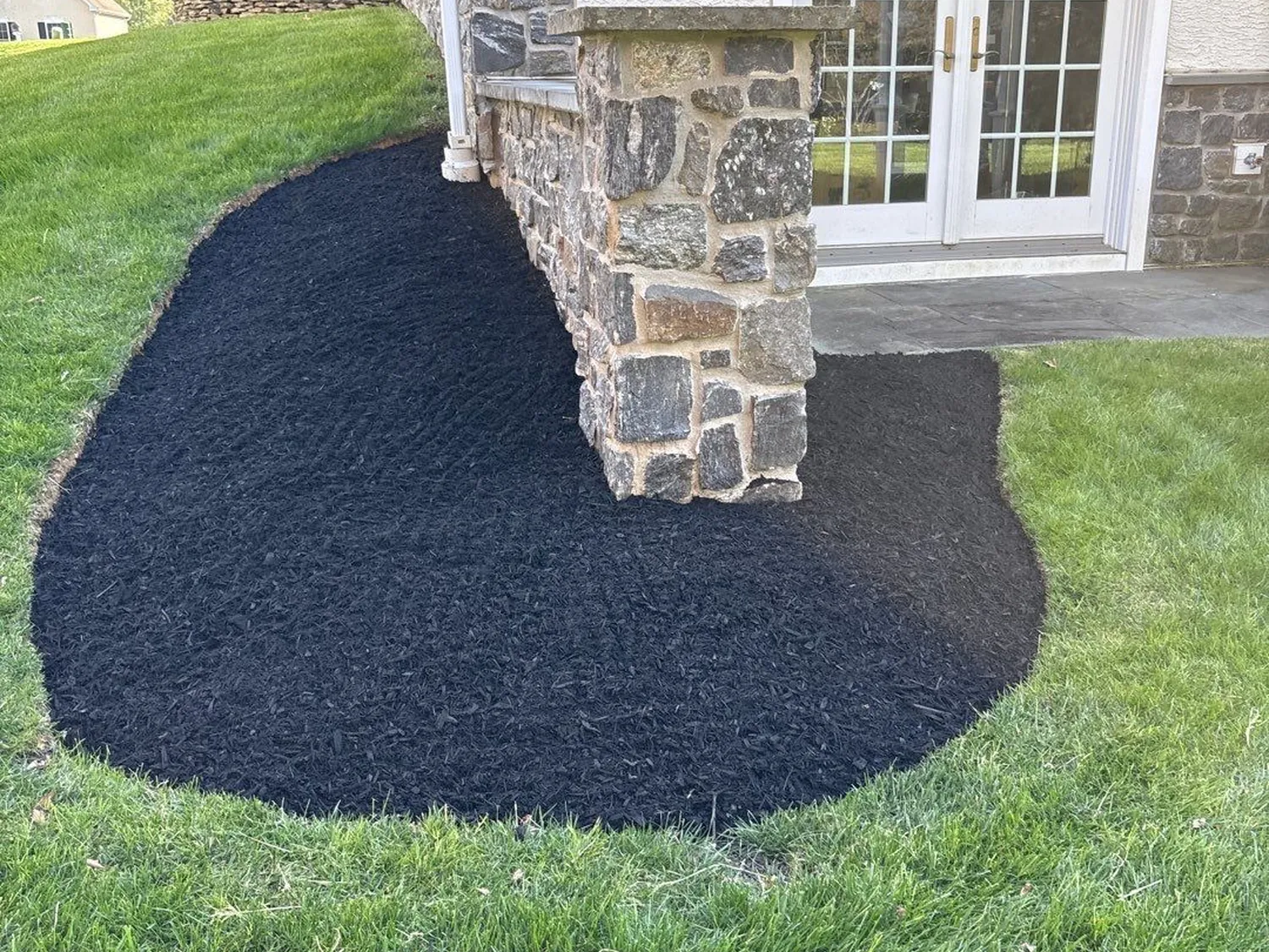 Freshly installed black shredded mulch bed wrapping the corner of a stone-faced home with French doors